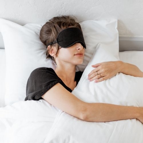Femme qui dort dans un lit blanc avec un masque noir sur les yeux en enlaçant un coussin